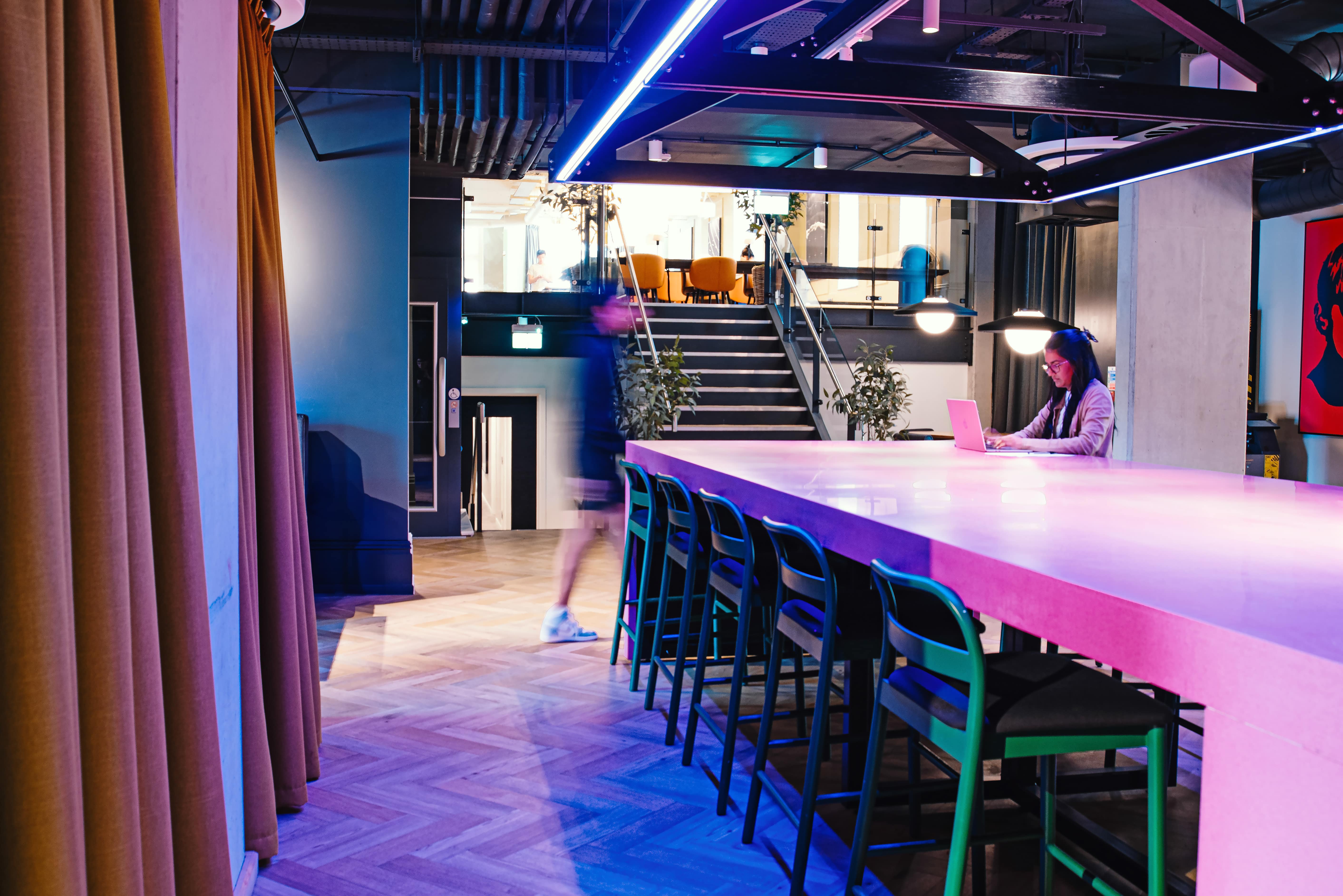 A modern office space with a long pink table surrounded by green chairs. A person sits at the end of the table using a laptop. Another person walks by, appearing blurred. Stairs and plants are visible in the background under bright lighting.